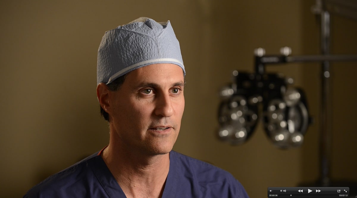 A physician in scrubs and surgical cap mid-sentence during a filmed interview, an eye-examination instrument behind him.